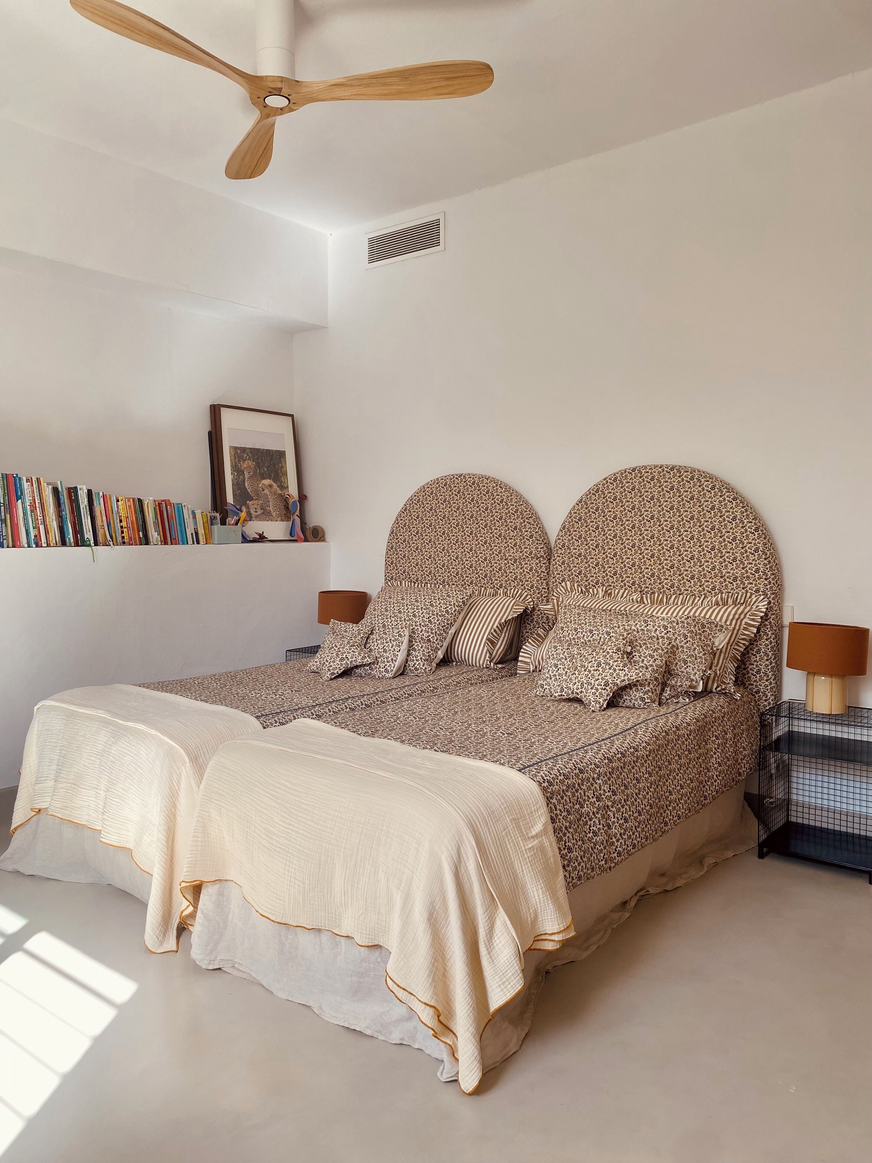 A bedroom with a bed, a ceiling fan, a wall-mounted shelf with books, and a framed picture.