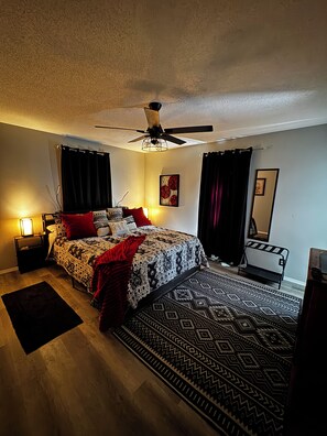 A bedroom with a bed, a ceiling fan, a nightstand, a bedside table, a rug, and a mirror.