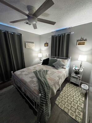 Un dormitorio con cama, dos mesitas de noche con lámparas, un ventilador de techo, una alfombra y cortinas.