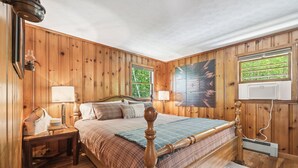A wooden-paneled bedroom with a bed, two bedside lamps, a nightstand, and a window with a view of trees.