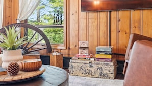 A cozy room with wooden walls, a large window with curtains, a wooden wheel, a vintage suitcase, and a stack of books.