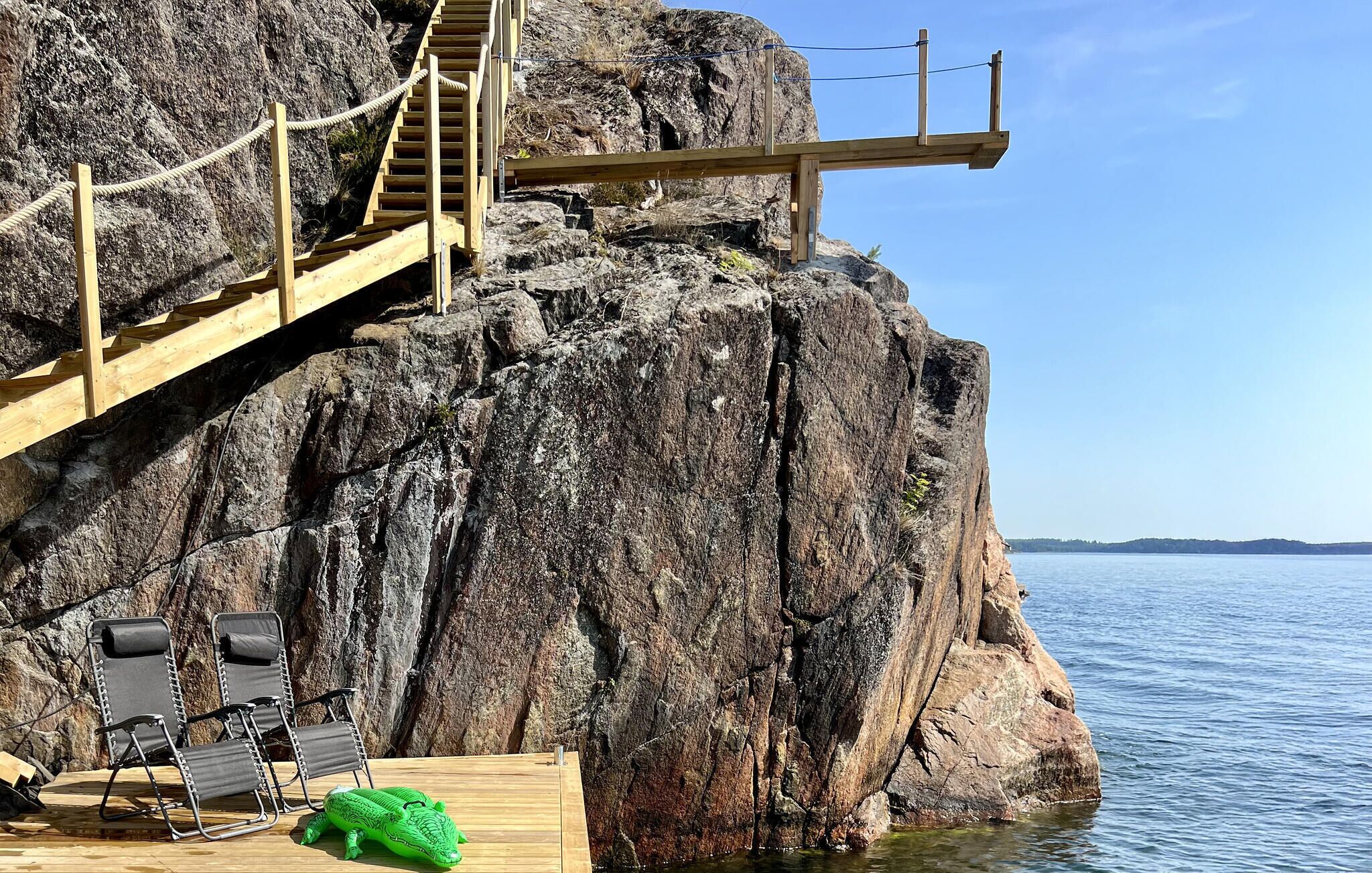 A wooden deck with a staircase leading up a rocky cliff, overlooking a body of water.