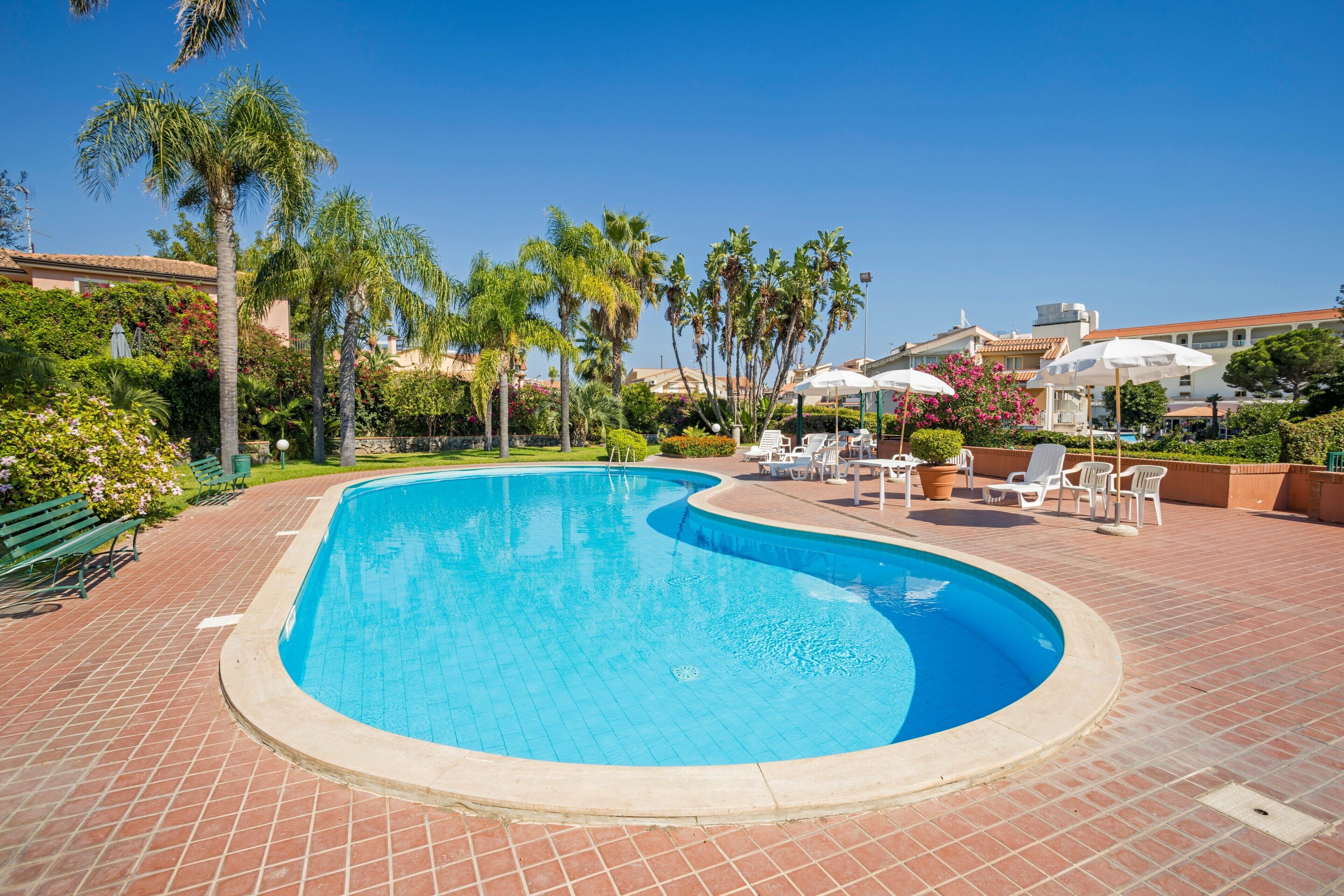 A swimming pool with a curved edge, surrounded by a brick patio, palm trees, and outdoor seating.