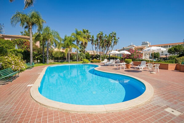 A swimming pool with a curved edge, surrounded by a brick patio, palm trees, and outdoor seating.