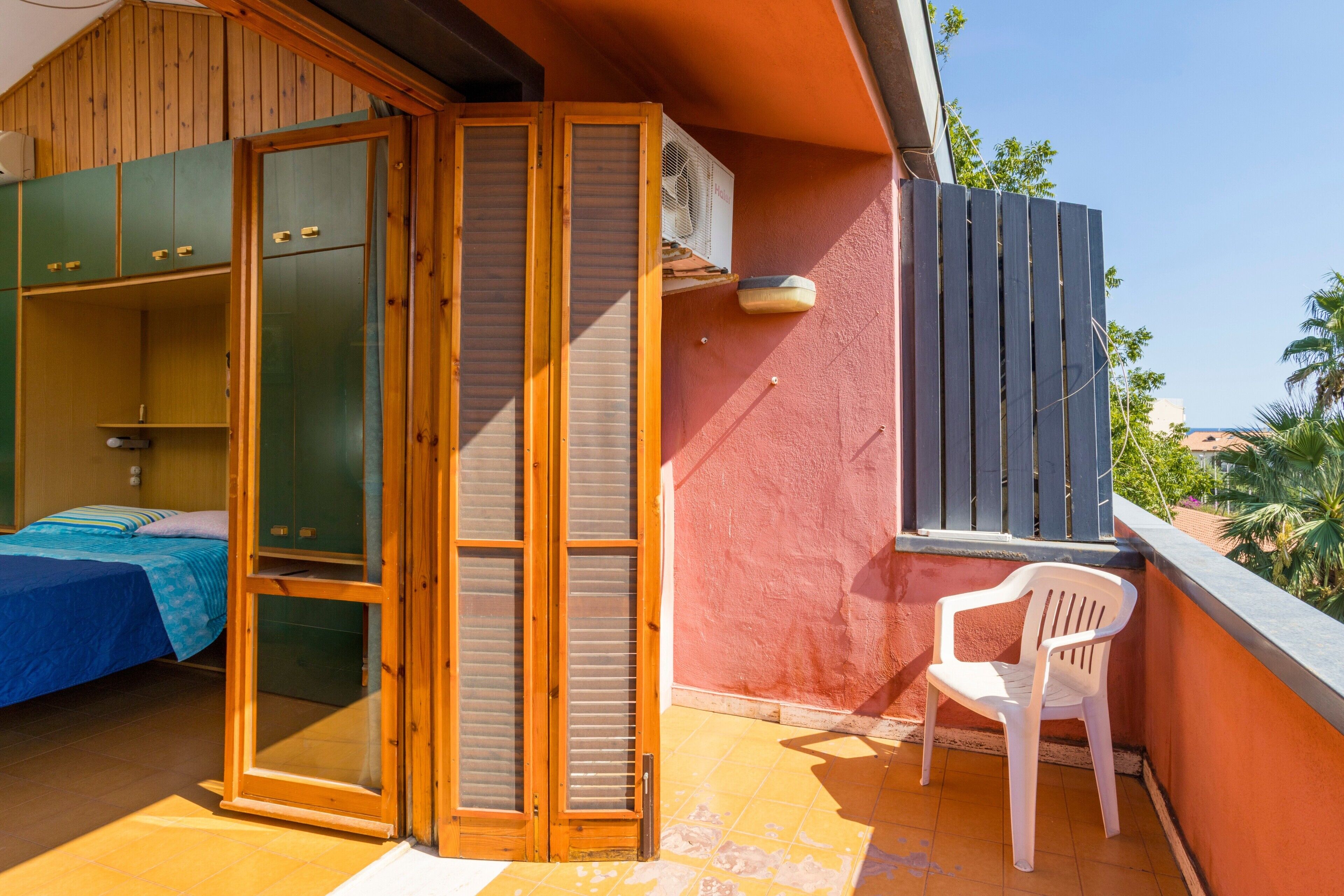 A room with a bed, a wooden wardrobe, a sliding glass door, and a balcony with a chair.