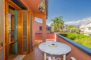 A balcony with a round table, wooden shutters, and a view of palm trees and other buildings.