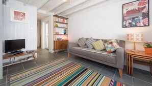 Ein Wohnzimmer mit grauem Sofa, farbenfrohem StreifenTeppich, Holznachttisch und einem Fernseher auf einem Holzboden.