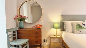 Ein Schlafzimmer mit einem Bett, einem Holzkasten, einem Sessel, einem Nachttisch mit Lampe, einem Spiegel mit Lichterkette und einer Vase mit Blumen.