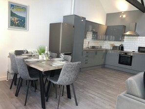 A modern kitchen with grey cabinets, a built-in oven, and a dining area with a wooden table and grey upholstered chairs. - Stunning Reverse Level Townhouse in Penzance (Penzance)