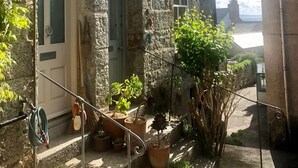 A stone building with a door, windows, and steps leading to a garden area with potted plants and a hose. - Janies Cottage- Eclectic Interiors & Vintage charm (Mousehole)