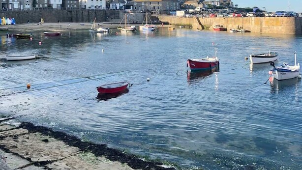 A coastal town with a harbor filled with boats, a stone wall, and buildings in the background. - Janies Cottage- Eclectic Interiors & Vintage charm (Mousehole)