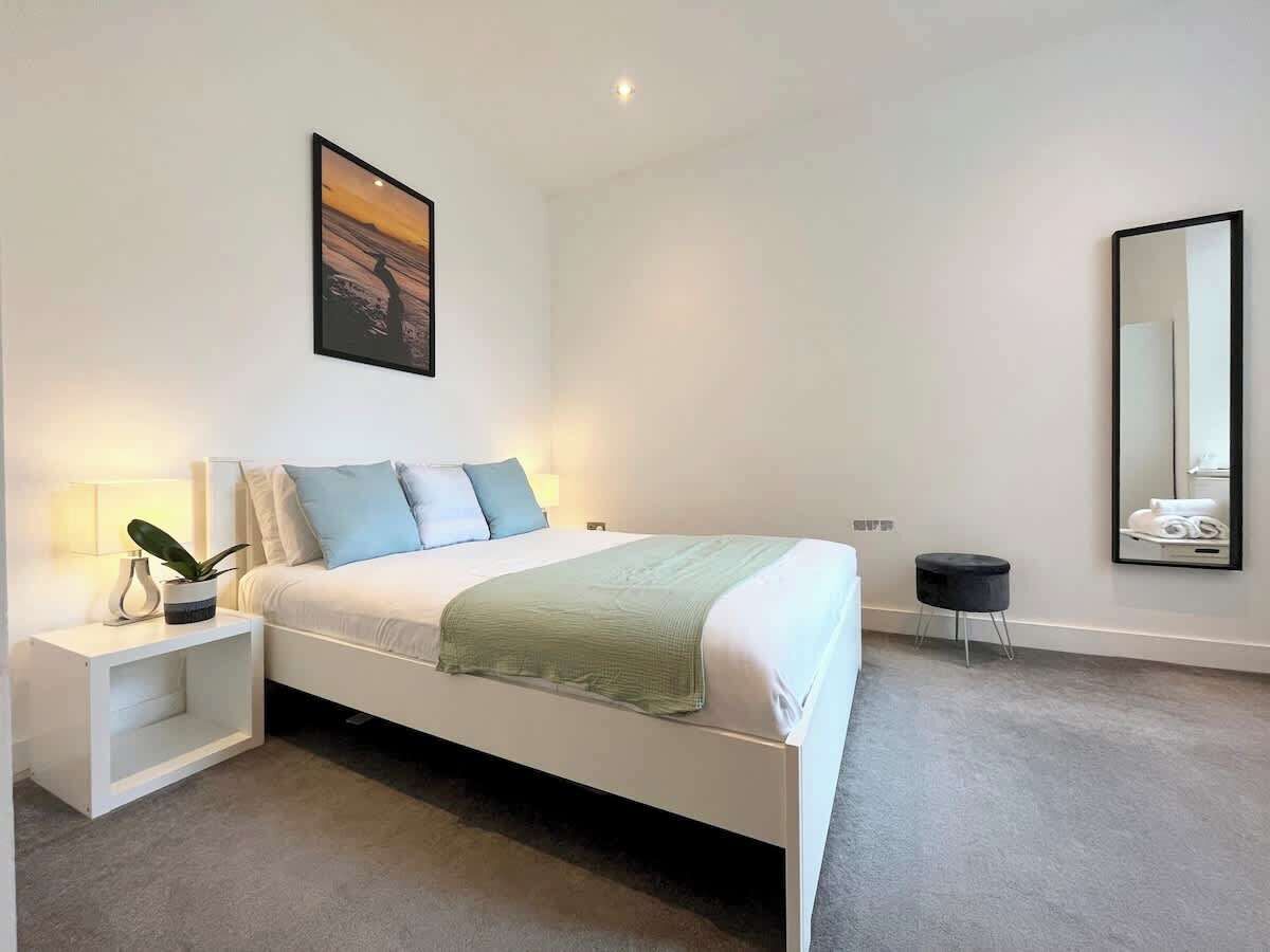 A modern bedroom with a white bed, a nightstand with a lamp, a small stool, and a wall-mounted mirror.