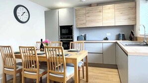A modern kitchen with wooden cabinets, a dining table with chairs, a clock on the wall, and a small island with a sink. - Kerensa, 3 bed with parking,  close to the beach (Penzance)