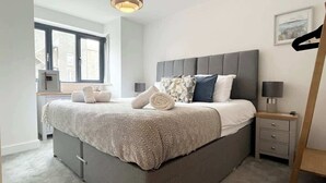 Un dormitorio moderno con una cama gris, una mesita de noche, una lámpara y una ventana con vistas a edificios.