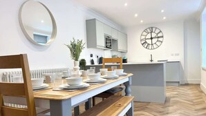 Une cuisine moderne avec des meubles blancs, une table à manger en bois mise avec des assiettes et des verres, une horloge au mur et un sol à motifs en chevrons.