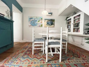 Et spiseområde med et hvidt bord og stole, en farverig måtte og et bogreol med bøger og planter.