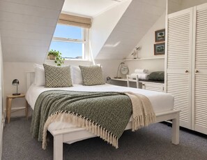 A bedroom with a sloped ceiling, a bed with patterned pillows, a bedside table with a lamp, a dresser, a mirror, and a view of the outdoors. - Charming 2-Bed Artist's Residence Central Penzance (Penzance)
