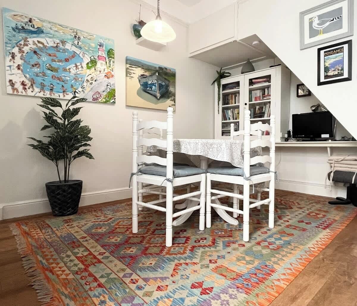 A dining area with a table and chairs, a patterned rug, a potted plant, and a bookshelf in the background.