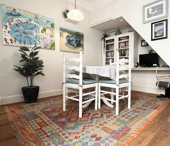 A dining area with a table and chairs, a patterned rug, a potted plant, and a bookshelf in the background.