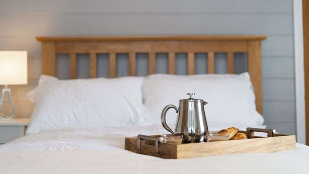 A neatly made bed with a wooden headboard, a tray with a coffee pot and pastries, and a bedside lamp.