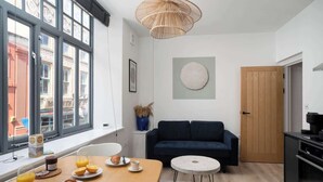 A modern living room with a large window, a dark blue sofa, a round coffee table, and a dining area with breakfast items. - Contemporary Apartment in central Penzance (Penzance)
