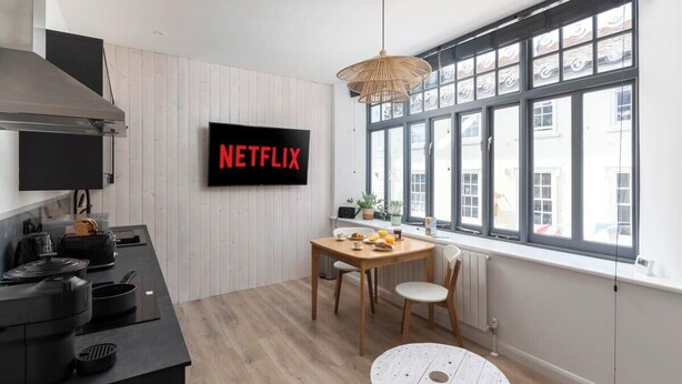 A modern kitchen with a large window, a dining table, and a TV mounted on the wall displaying "NETFLIX". - Contemporary Apartment in central Penzance (Penzance)