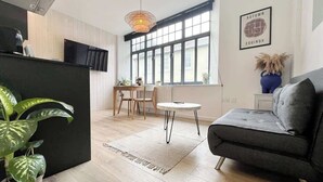 A modern living room with a sofa, a dining table, and chairs. There is a large window with a view of the outside. - Contemporary Apartment in central Penzance (Penzance)