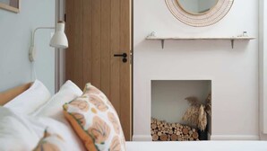 A bedroom with a bed, a wooden door, a wall-mounted lamp, a fireplace with logs and a plant, and a shelf with decorative items. - Contemporary Apartment in central Penzance (Penzance)
