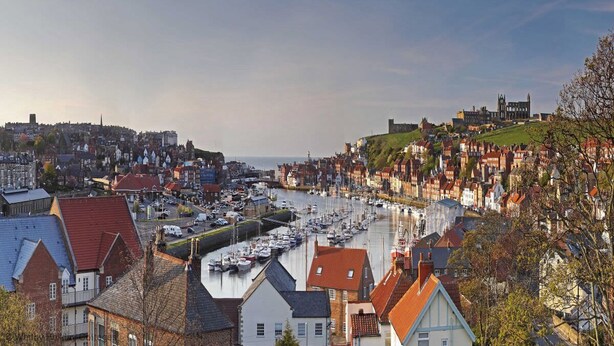 A coastal town with a marina filled with boats, historic buildings, and a prominent church on a hill.