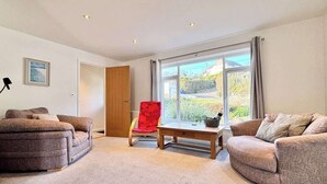 Ein geräumiges Wohnzimmer mit zwei Sofas, einem roten Sessel, einem Couchtisch und einem großen Fenster mit Blick ins Freie.