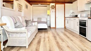A cozy room with a wicker sofa, a bed, a wooden ceiling, and a kitchen area with white cabinets and appliances.