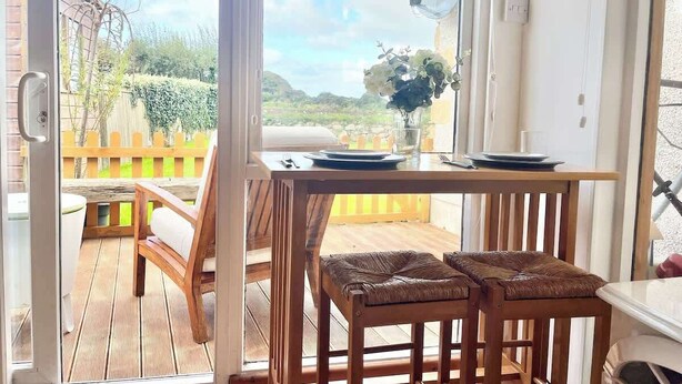 A wooden deck with a table and two wicker chairs, a wooden fence, and a view of a garden and hills in the background.