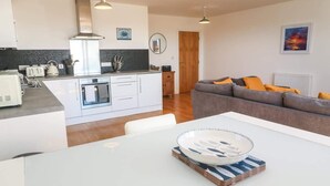 Una cocina moderna con armarios blancos, una campana extractora de acero inoxidable y un protector contra salpicaduras negro. Hay un comedor con una mesa blanca y un plato con diseño azul y blanco. Un sofá gris con cojines naranjas se encuentra en la sala de estar.