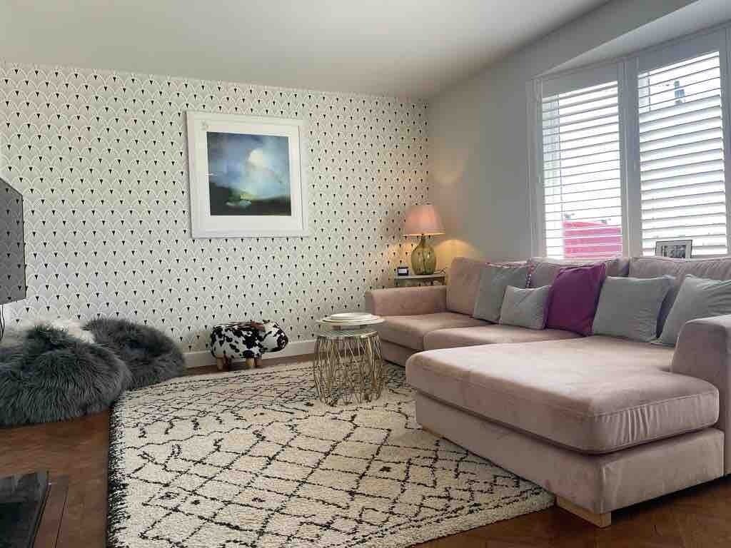 Una sala de estar con un papel tapiz estampado, un sofá, una alfombra, una lámpara y un cuadro enmarcado.