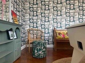A room with a patterned wallpaper, a wooden chair, a green ottoman, a bed with a colorful pillow, and a cabinet with framed pictures.