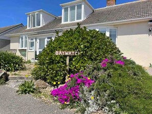 A house with a well-maintained garden, a sign reading 'BAYWATCH,' and a clear blue sky.