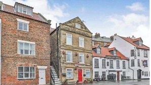 A row of traditional brick buildings with a red door, white windows, and a pub sign. - Waterfront Cottage by Whitby Harbour + (Whitby)