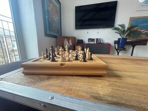A chessboard on a table with a window view, a television, and a bookshelf in the background. - Stylish~Tasteful~2Bed~2Bath~Central~Townhouse (Penzance)