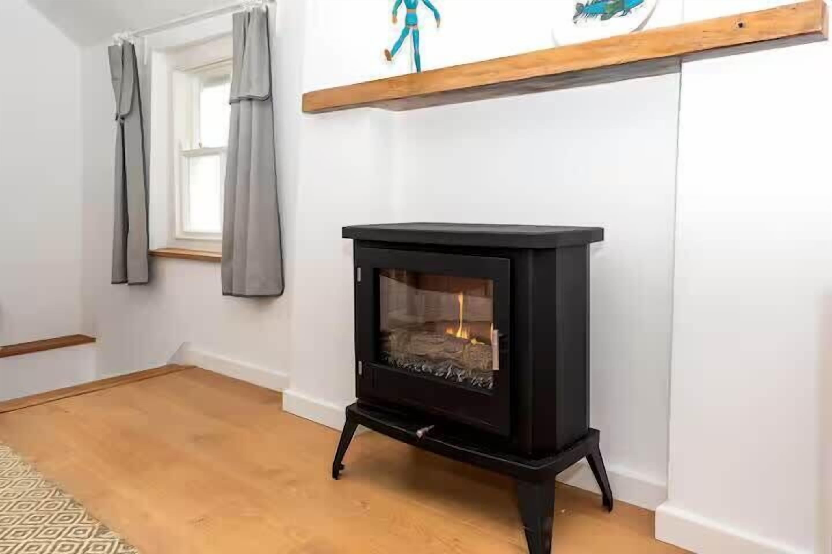 Une pièce avec un poêle à bois noir, une fenêtre ornée de rideaux gris et une étagère en bois au-dessus du poêle.