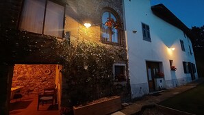 Un edificio in pietra con una panca e un tavolo in legno all'esterno, un edificio bianco con una finestra e una fioriera, e un ingresso ben illuminato.