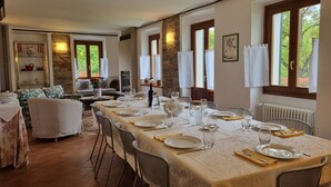 Zona pranzo con un tavolo lungo apparecchiato per un pasto, circondato da sedie in vimini e ampie finestre con tende.