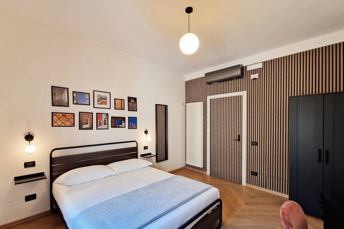 Une chambre à coucher moderne, comprenant un lit, des tables de chevet, des œuvres d’art accrochées au mur, une armoire et un luminaire suspendu.