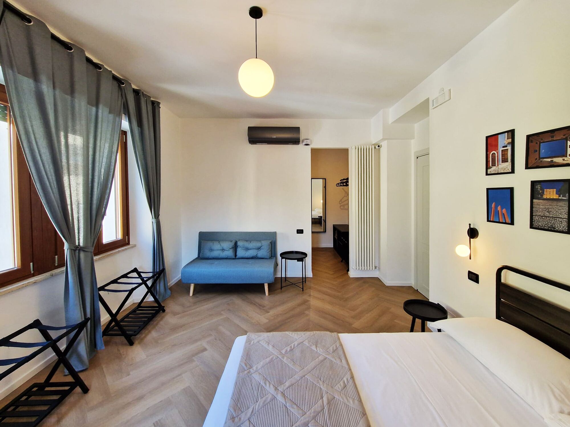 Een moderne hotelkamer met een groot bed, een bank, een kleine tafel en een stoel. Er is een raam met gordijnen, een radiator en wandlampen.
