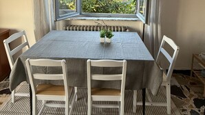 Un'area pranzo con un tavolo ricoperto da una tovaglia grigia, circondato da quattro sedie bianche. Sul tavolo ci sono due piante in vaso.