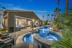 A pool area with lounge chairs and a fire pit, surrounded by palm trees and a building with a tiled roof. - Lotus Retreat: 7BD Pool Game Rm (Indio)