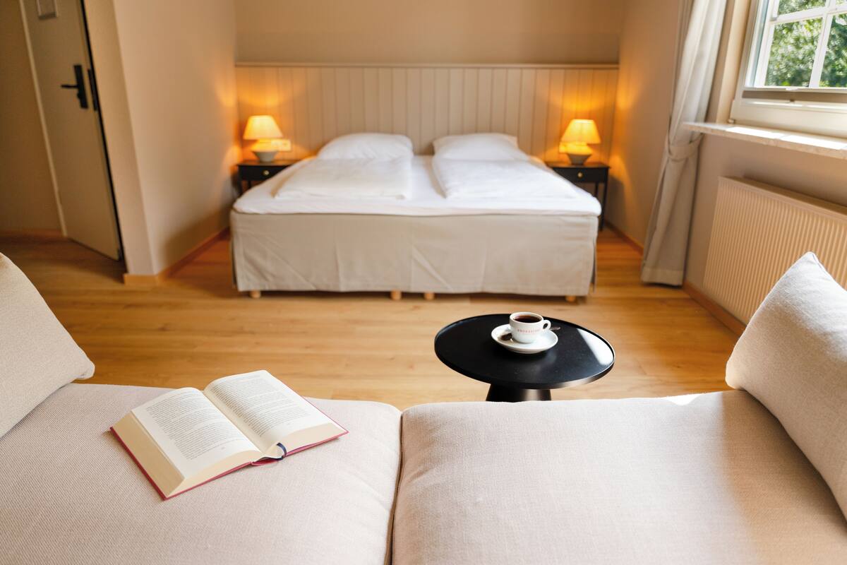 A bedroom with a double bed, bedside lamps, a small table with a cup, and an open book on a sofa.