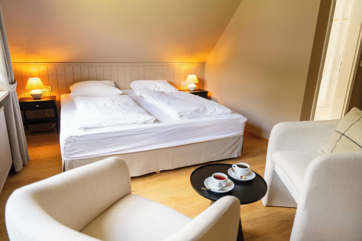 A hotel room with a double bed, two bedside tables with lamps, a small round table with a cup of coffee, and two armchairs.