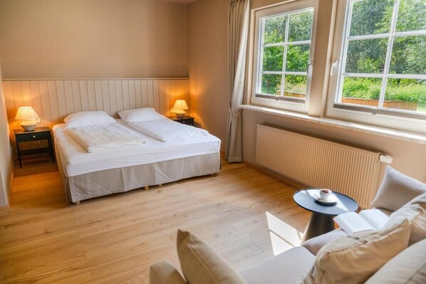 A hotel room with a double bed, bedside lamps, a small table with a cup, and a view of greenery through the window.