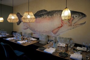 A restaurant dining area with tables set for service, a large fish mural on the wall, and hanging wicker lamps.