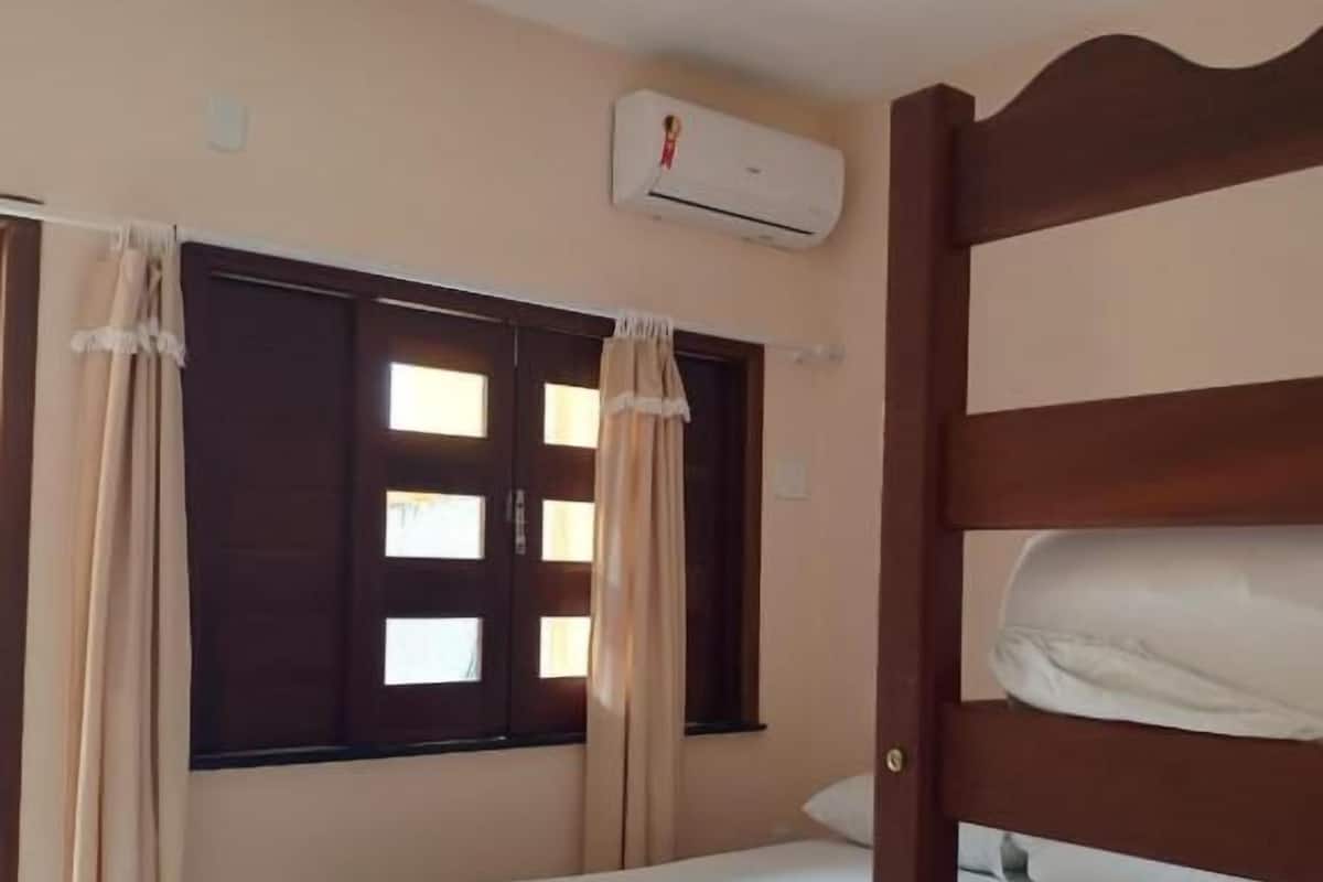 Uma cama de solteiro com cabeceira de madeira, cortinas bege e um aparelho de ar condicionado na parede.